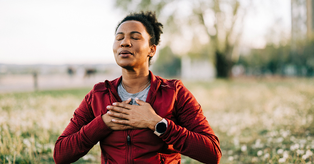 Woman outdoors, meditating with eyes closed and hands on chest, enhanced wellbeing and health.